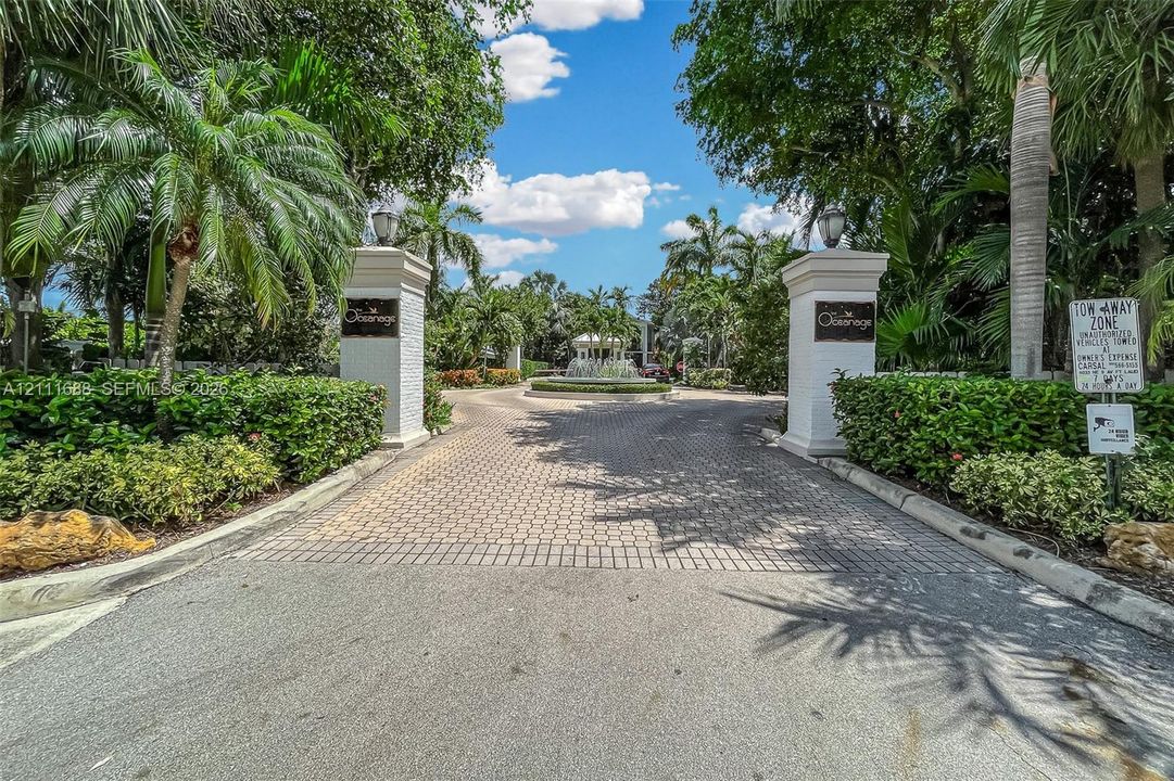 Gated entrance featuring decorative stone pillars, paver driveway, mature palm trees, lush perimeter landscaping, and a central fountain