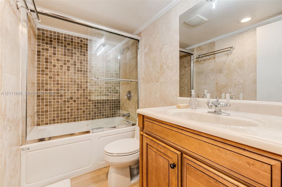 Bathroom featuring stone wall tiling, wood-finish vanity with an integrated countertop sink, mosaic tile shower surround, glass shower enclosure, and chrome faucet hardware