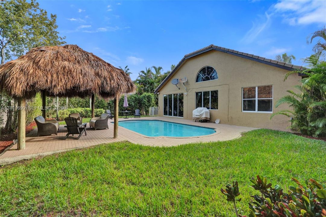 Stucco-exterior residence featuring an in-ground swimming pool, a thatched-roof tiki hut, a paved patio, and a manicured lawn