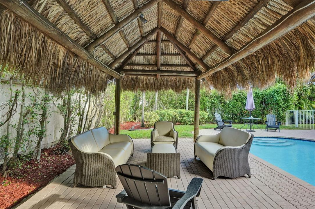 Outdoor thatched-roof pavilion featuring a timber-post structure and paver flooring, situated on a poolside patio