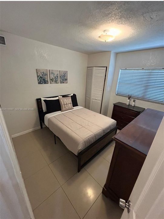 Room featuring neutral tile flooring, window with horizontal blinds, ceiling-mounted light fixture, and closet with louvered doors