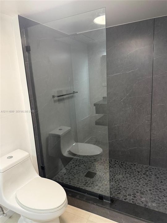 Walk-in shower featuring a frameless glass enclosure, floor-to-ceiling stone-patterned tile surround, pebble-style stone flooring, and a recessed shower head