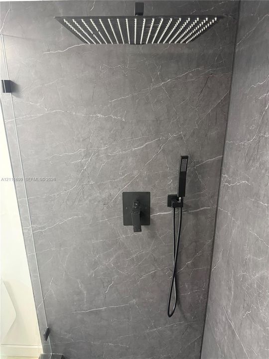 Shower enclosure featuring grey stone-finish tile surround, oversized square rainfall showerhead, matte black handheld spray, and coordinated wall-mounted control hardware