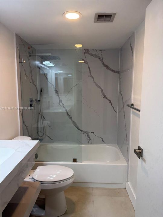Bathroom featuring a white vanity with a composite countertop, a porcelain-finish bathtub, and a full-height wall surround with a veined pattern behind a frameless glass shower panel