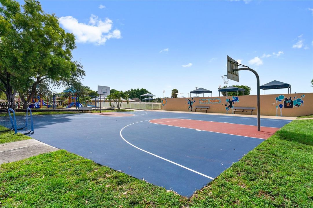 Outdoor multi-sport court featuring a paved basketball surface, metal backboards, perimeter bench seating, and shaded structures