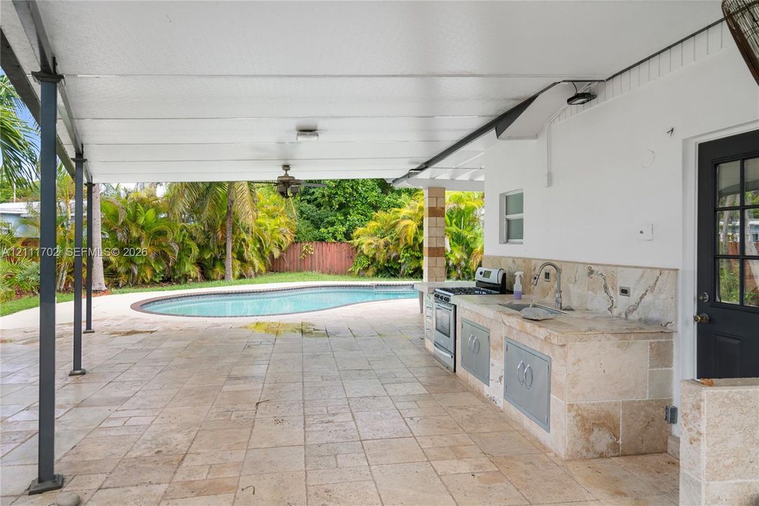 Covered patio featuring natural stone tile flooring, an outdoor kitchen with integrated stone-clad cabinetry, a stainless steel range, and a sink, overlooking a swimming pool