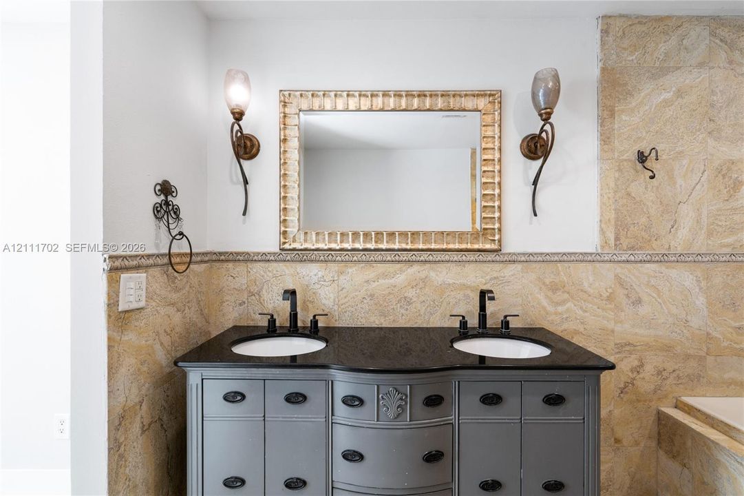 Dual-sink vanity with dark stone countertop, integrated undermount sinks, custom-finish cabinetry, ornate metallic wall sconces, and neutral stone tile wall surrounds