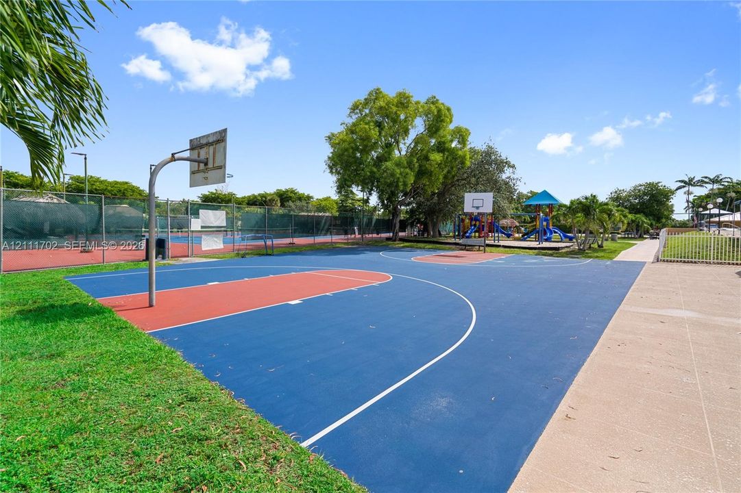 Multi-purpose recreational area featuring a paved basketball court, fenced tennis court, and outdoor playground equipment