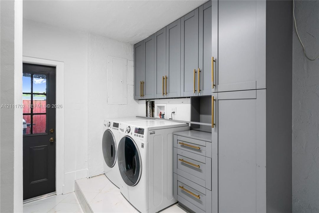 Laundry area featuring grey Shaker-style cabinetry, matte gold-finish hardware, and a dark-toned exterior door with glass pane detail