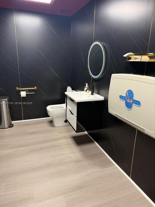 Modern bathroom featuring black wall paneling with gold-toned trim, a floating vanity with a white basin, a wall-mounted illuminated circular mirror, and wood-finish flooring