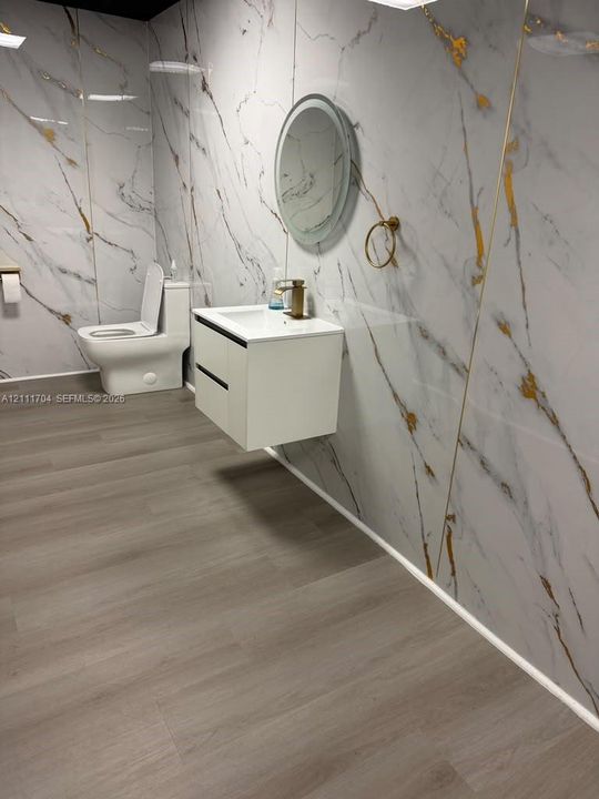 Large-format stone-pattern wall panels with gold veining, floating white dual-drawer vanity, wall-mounted round backlit mirror, gold-tone hardware, and wood-finish flooring