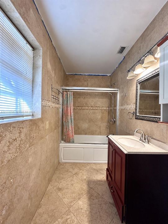 Full-height stone wall tiling with decorative inlay, walk-in bathtub with sliding glass doors, dark wood-finish vanity, light-toned stone countertop with an integrated sink, and wall-mounted vanity lighting