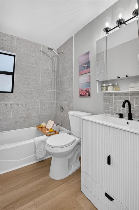 Bathroom featuring a ribbed-detail vanity with a matte black faucet, wood-finish flooring, a tub-shower combination with full-height stone-look tile surround, and a tri-light vanity fixture