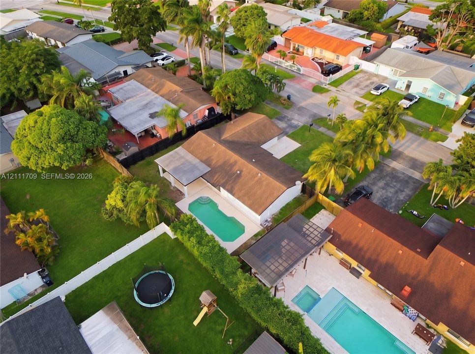 Aerial perspective of a single-story residence featuring a shingled roof, a covered patio, and an in-ground swimming pool situated on a fenced lot with mature landscaping