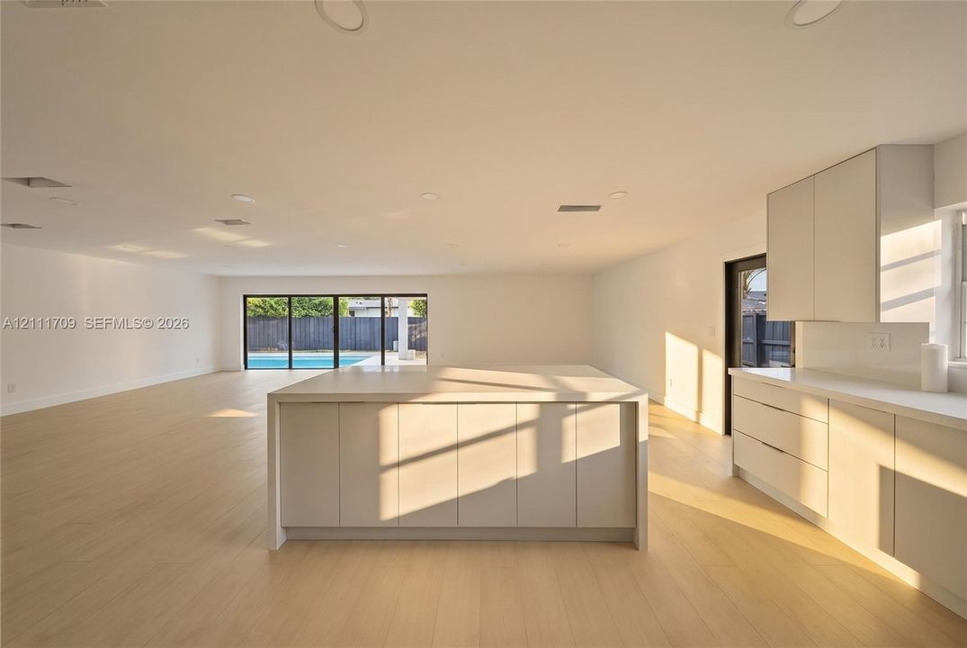 Open-concept interior featuring light-toned wood-finish flooring, a central kitchen island with integrated cabinetry, matte finish flat-panel cabinetry, and multi-panel sliding glass doors