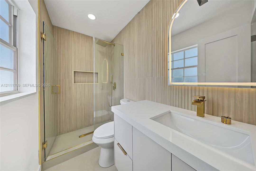 Bathroom featuring white vanity with integrated basin, gold-toned faucet, arched LED vanity mirror, vertical textured wall tile, and walk-in shower with glass enclosure