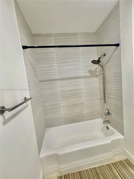 Full-height wall surround with textured, linear-patterned tile, white porcelain soaking tub, chrome handheld shower fixture, and brushed metal towel bar