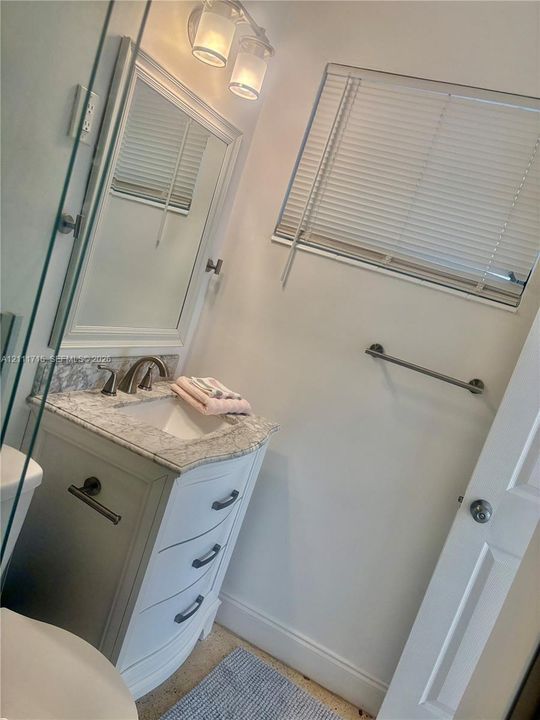 White vanity featuring a stone-composite countertop with an integrated basin, three-drawer storage, framed vanity mirror, dual-bulb wall-mounted lighting, and a horizontal window with adjustable blinds