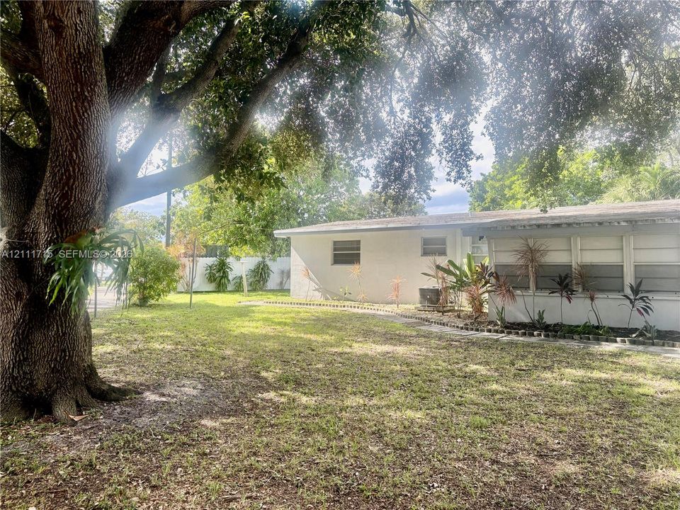Ground-level exterior featuring a painted masonry facade, mature shade trees, a stone-bordered garden bed, and a glass-paneled sunroom