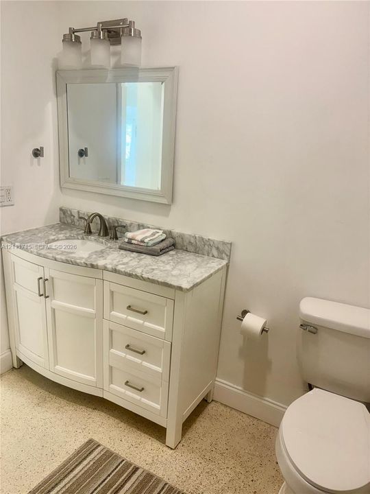 White vanity cabinet with drawers, light-toned stone-finish countertop, undermount sink, three-light wall fixture, and framed wall mirror