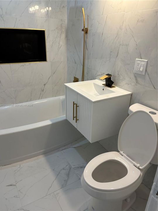 Wall-mounted white vanity with a rectangular sink, gold-tone hardware, full-height stone-patterned wall tiling, soaking tub, and a gold-tone handheld shower fixture
