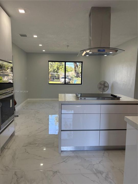 Modern kitchen featuring white flat-panel cabinetry, a central island with a cooktop, stone-finish flooring, a stainless steel chimney-style range hood, and built-in wall oven