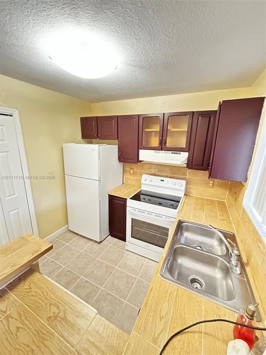 Kitchen featuring dark-toned cabinetry, tile countertops, tile backsplash, built-in dual-basin stainless steel sink, and white appliances