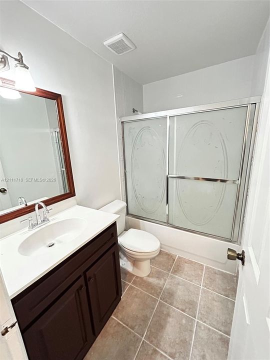 Dark wood-finish vanity with a white countertop and oval undermount sink, framed wall mirror, tiled flooring, and a shower enclosure featuring etched glass sliding doors
