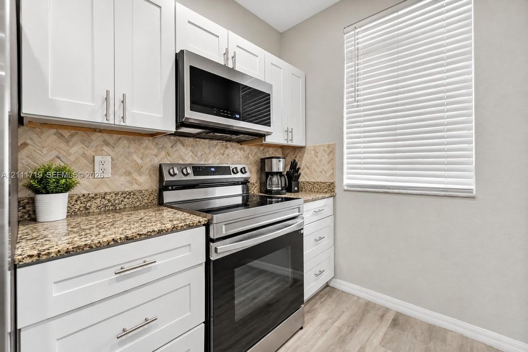 Kitchen featuring white shaker-style cabinetry, stone-finish countertops, stainless steel appliances, a herringbone-patterned tile backsplash, and wood-finish flooring