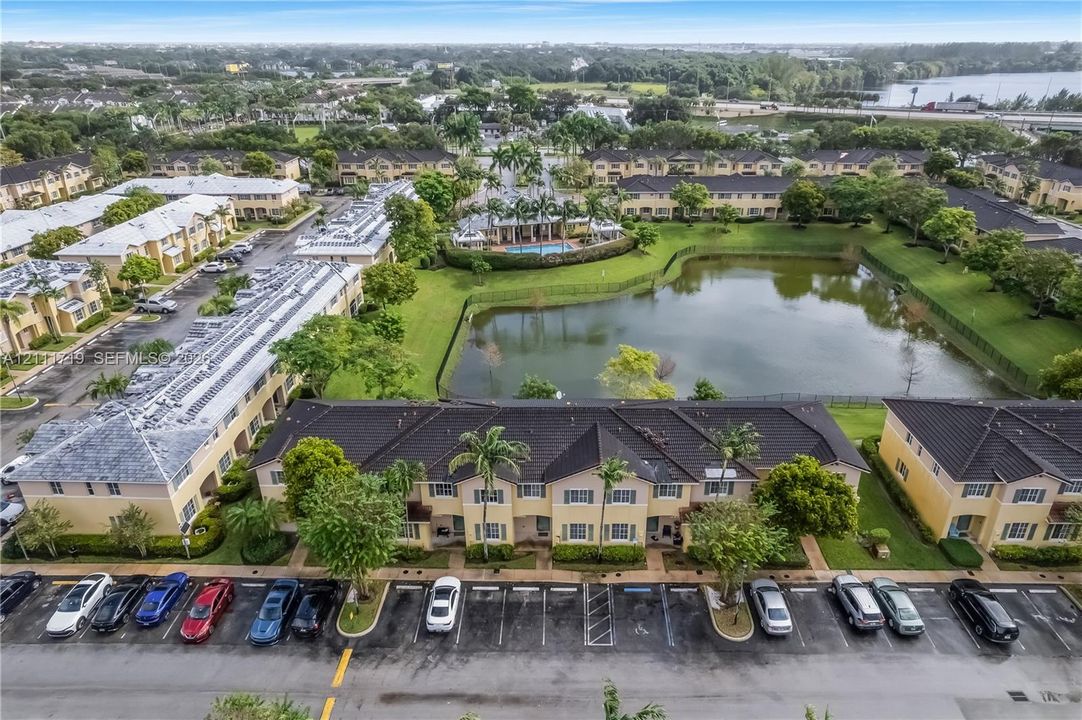 Aerial perspective featuring multi-story residential structures with tile roofing, surface parking, and a community pool area centered around a pond