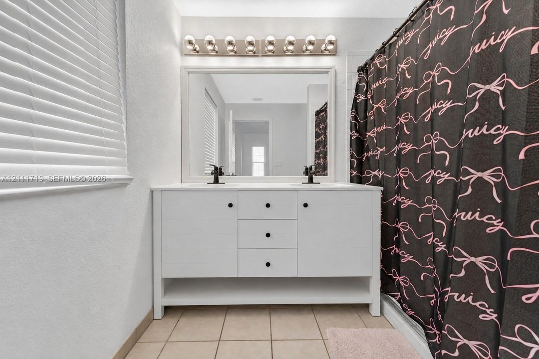 White dual-sink vanity with central drawer storage, large framed mirror, multi-bulb wall-mounted lighting, and ceramic tile flooring