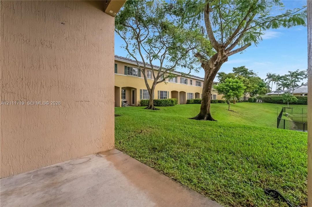 Covered concrete patio with stucco exterior, expansive lawn, mature trees, and metal perimeter fencing