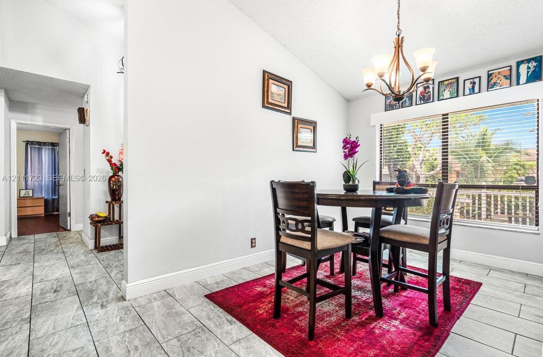 Vaulted ceilings, large window with horizontal blinds, and tile flooring featuring stone-finish detailing, illuminated by a multi-arm chandelier