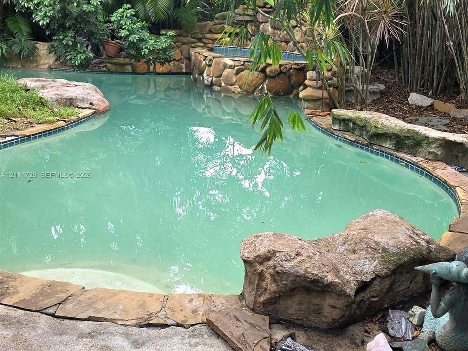 Freeform swimming pool featuring a natural stone border, integrated stone waterfall, built-in tiled spa, and large decorative boulders