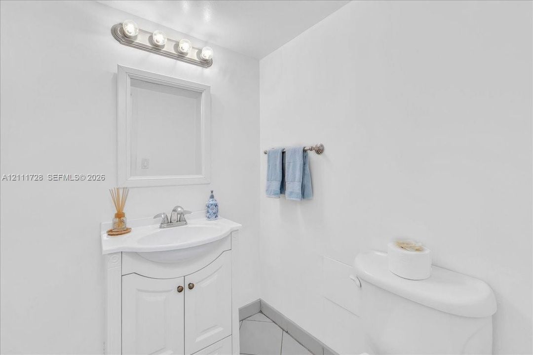 White vanity with a molded countertop and integrated basin, framed wall mirror, four-light vanity fixture, and neutral tile flooring