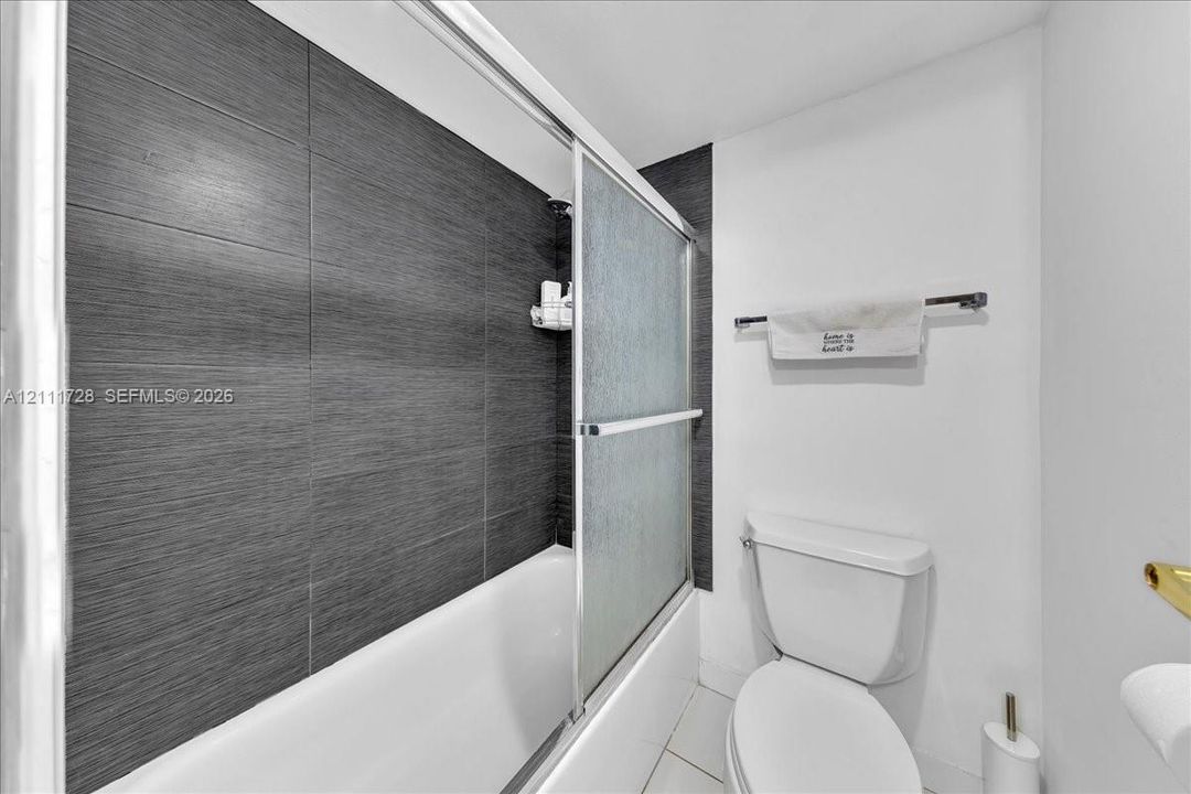Bathroom featuring large-format dark grey textured tile shower surround, sliding glass shower door, white acrylic tub, and elongated porcelain toilet