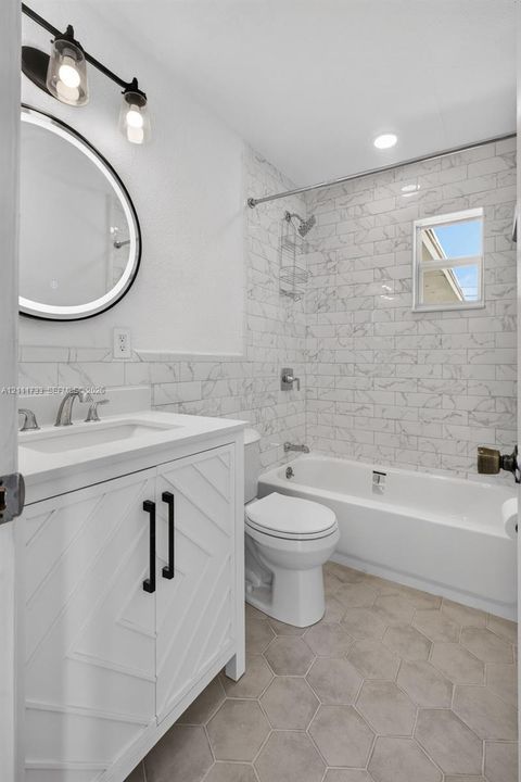 Main Bathroom featuring a white chevron-pattern vanity with a solid-surface countertop, an illuminated round mirror, porcelain tile shower surround, and hexagonal floor tile