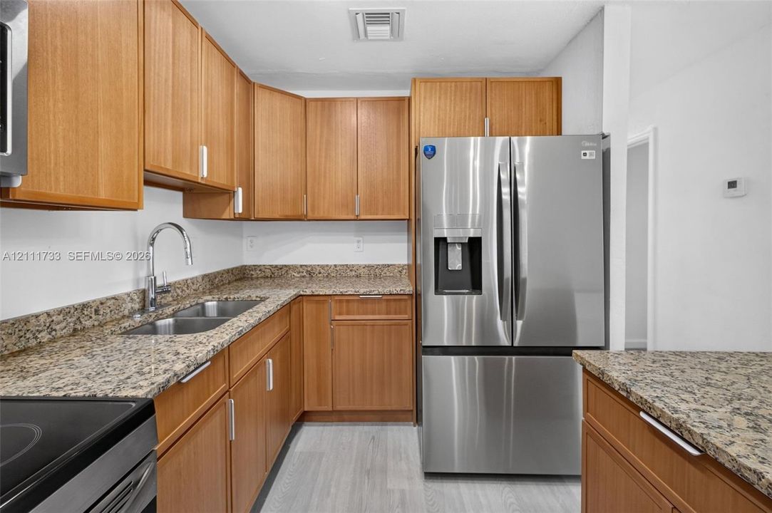 Kitchen featuring wood-finish cabinetry, stone countertops, stainless steel refrigerator, undermount double-basin sink with gooseneck faucet, and light-toned flooring