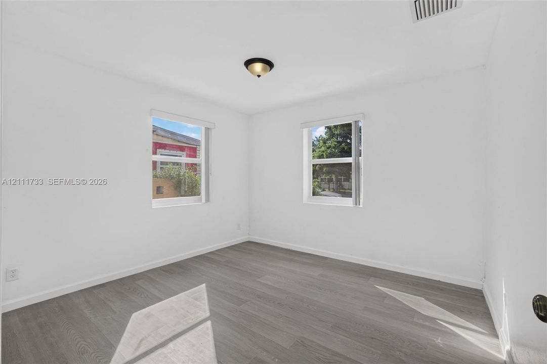 Larger Front bedroom - dual exterior view windows featuring white walls, wood-finish flooring, and windows with horizontal blinds, illuminated by a ceiling-mounted flush light fixture
