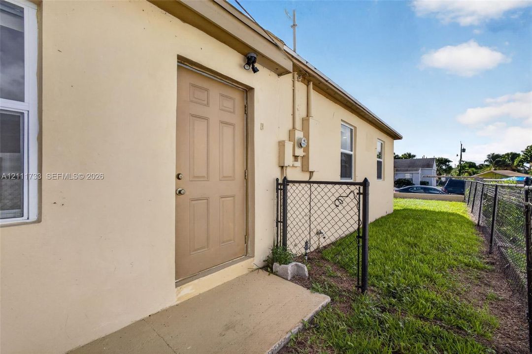 Stucco exterior featuring a solid panel door, exterior wall-mounted light fixture, concrete threshold, and chain-link fencing enclosing the side yard
