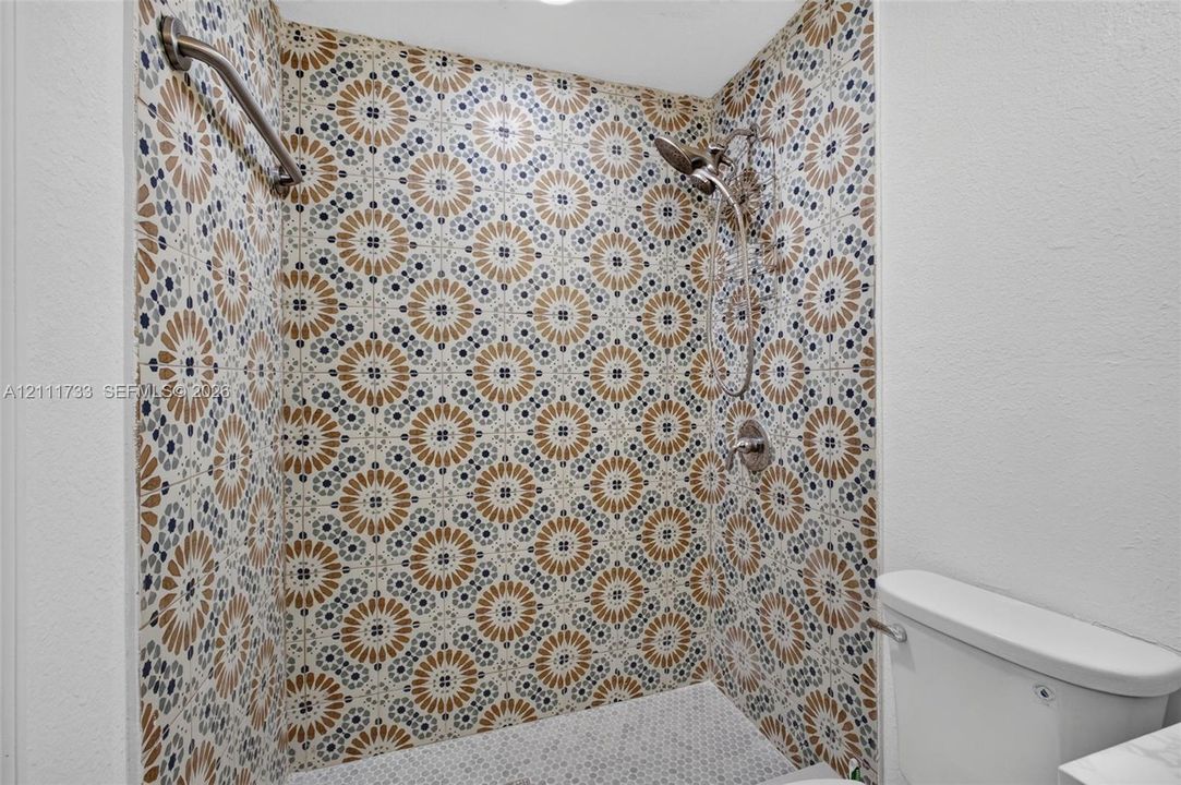 Walk-in shower featuring decorative patterned tile walls, a handheld showerhead, a metal grab bar, and a mosaic tile floor