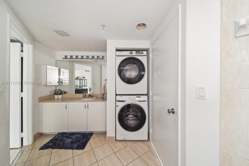 Built-in laundry closet with stacked washer and dryer, vanity featuring solid surface countertop, integrated sink, and oversized vanity mirror with overhead theater-style lighting, finished with ceramic tile flooring