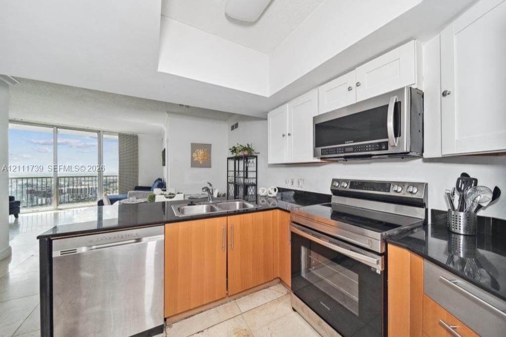 Kitchen featuring natural wood-finish lower cabinetry, white upper cabinets, black stone countertops, stainless steel appliances, and tile flooring