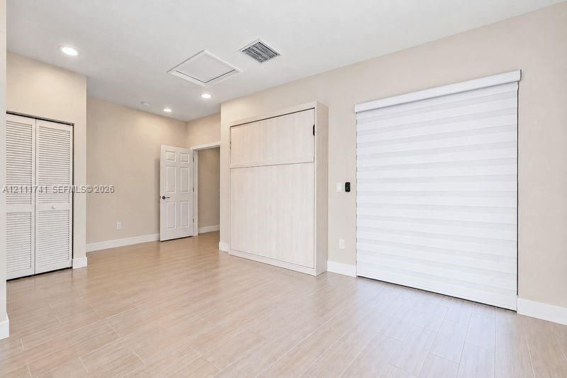 Neutral-toned interior featuring wood-finish tile flooring, recessed ceiling lighting, a built-in cabinet unit, dual louvered-door closet, and motorized roller shades