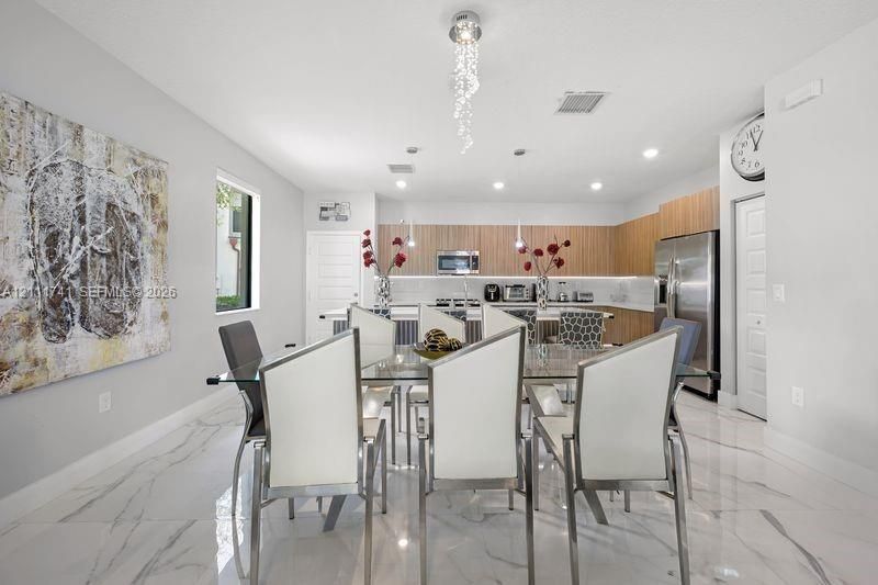 Open-concept kitchen and dining area featuring large-format tile flooring with marble-style veining, flat-panel wood-finish cabinetry, stainless steel appliances, and a central pendant light fixture with decorative glass elements