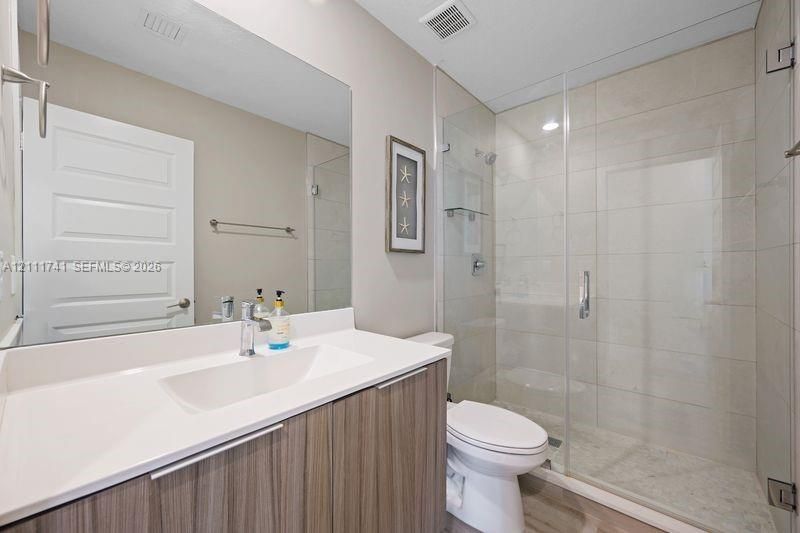 Bathroom featuring a vanity with a white countertop and integrated basin, large wall-mounted mirror, glass-enclosed shower with tiled surround, and wood-finish cabinetry