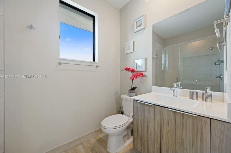 Bathroom featuring a wood-finish floating vanity, integrated rectangular sink, light-toned stone countertop, large wall-mounted mirror, and glass-enclosed shower with tile surround