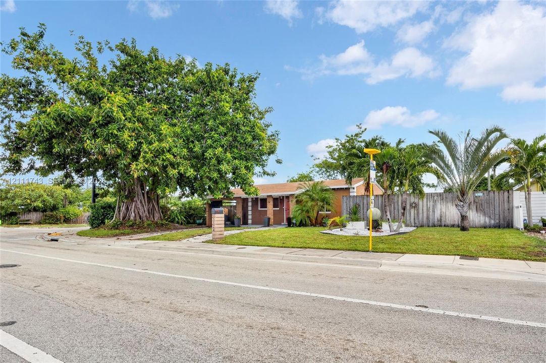 Single-story residence featuring a horizontal wood-slat privacy fence, manicured front lawn, mature specimen tree, and tropical landscaping