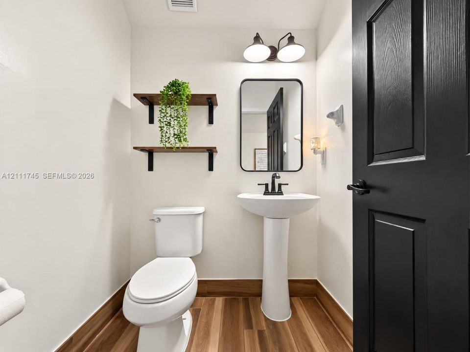 Powder room featuring a white pedestal sink with matte black hardware, rounded rectangular mirror, two-light fixture, wood-finish flooring, and floating wall-mounted shelving