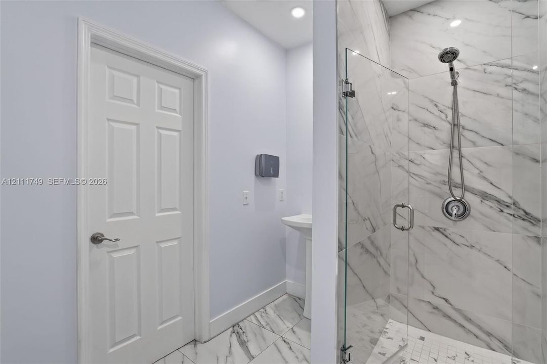 Walk-in shower featuring glass paneling, tile surrounds with veining, and a handheld shower fixture, complemented by pedestal sink and tile flooring